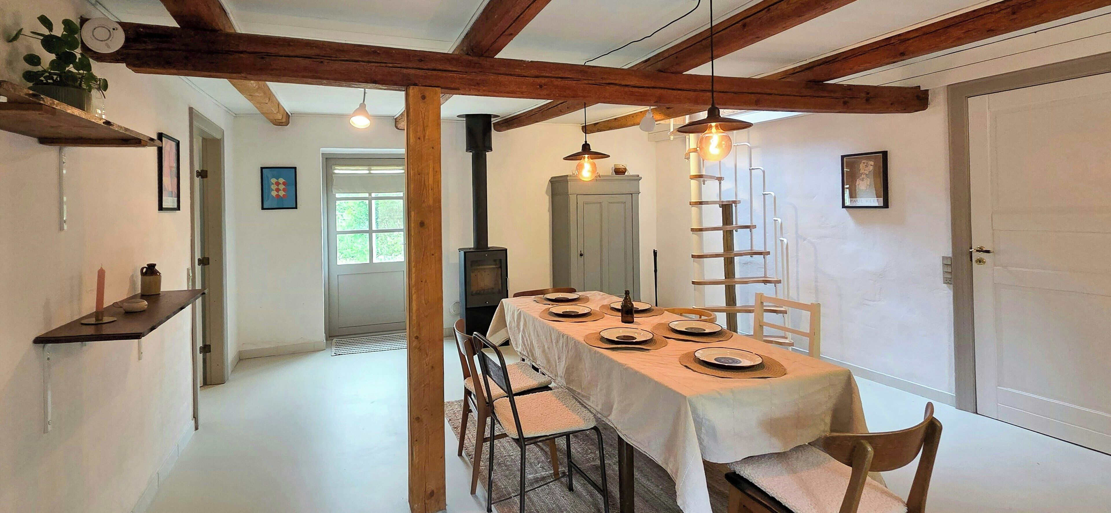 Un comedor con una mesa larga dispuesta para cuatro personas, sillas de madera, un mantel blanco y techo con vigas de madera.
