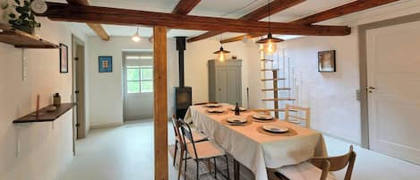 A dining area with a long table set for four, wooden chairs, a white tablecloth, and a wooden beam ceiling.