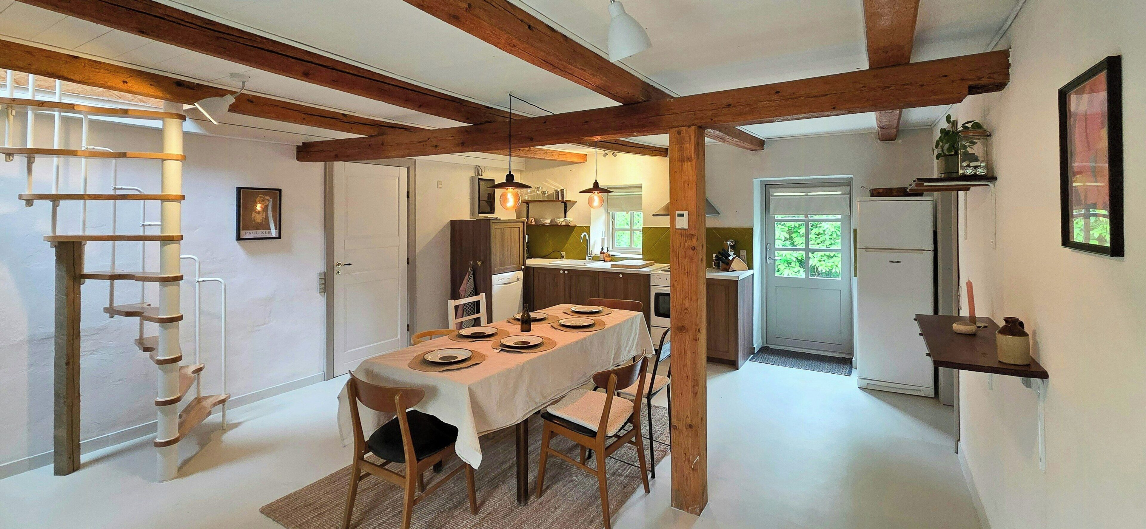 A dining area with a table set for four, wooden beams on the ceiling, a kitchen in the background, and a door leading outside.