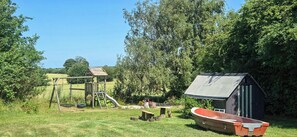A playground with a slide and swings, a small shed, and a boat in a grassy area with trees.