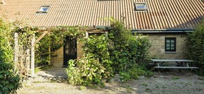 Een landelijk pand met een bakstenen gevel, een houten pergola met klimplanten en een grindpad dat naar een houten deur leidt.