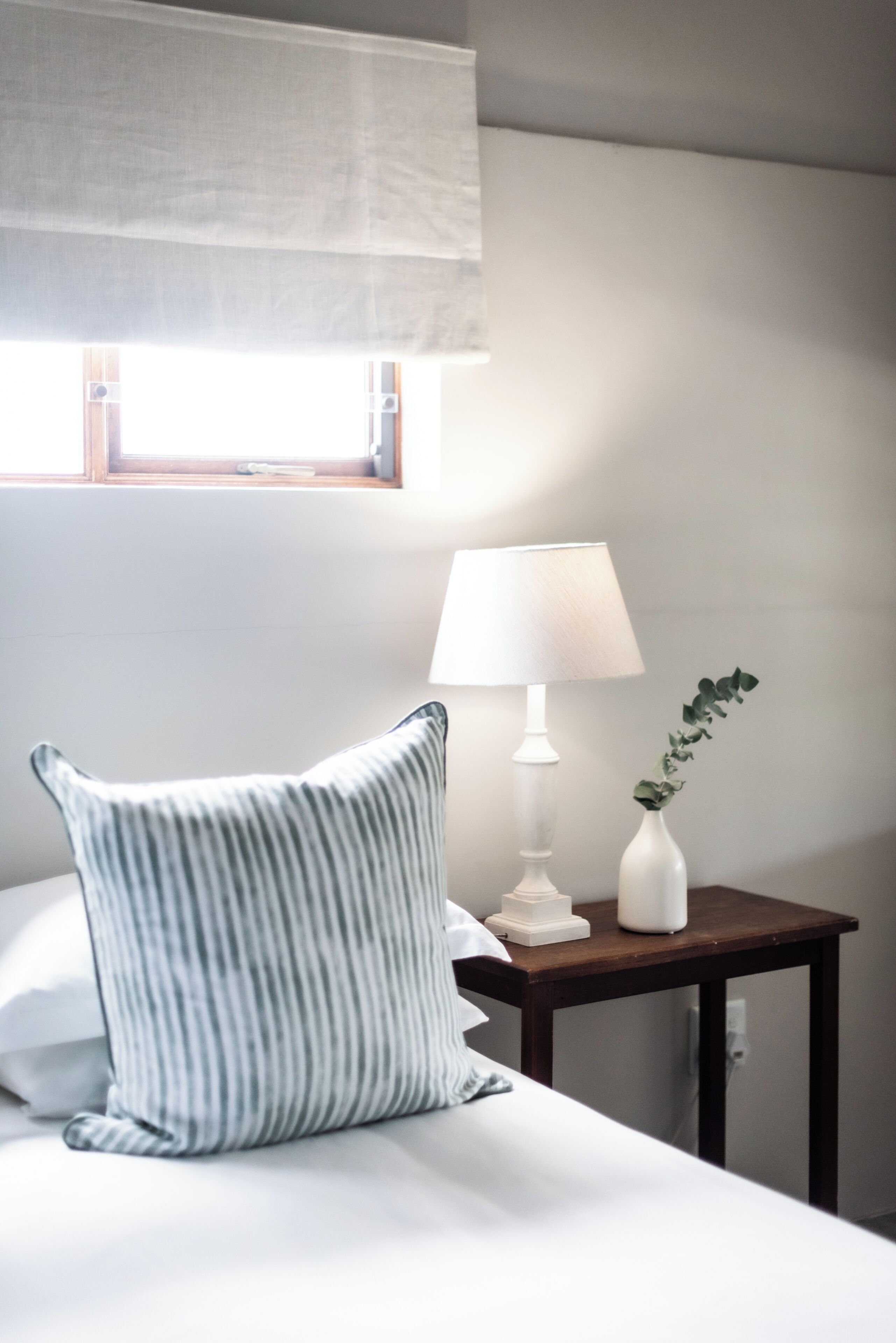 Et hotelværelse med en seng, et natbord med lampe, et vindue med rullegardin og en potteplante.