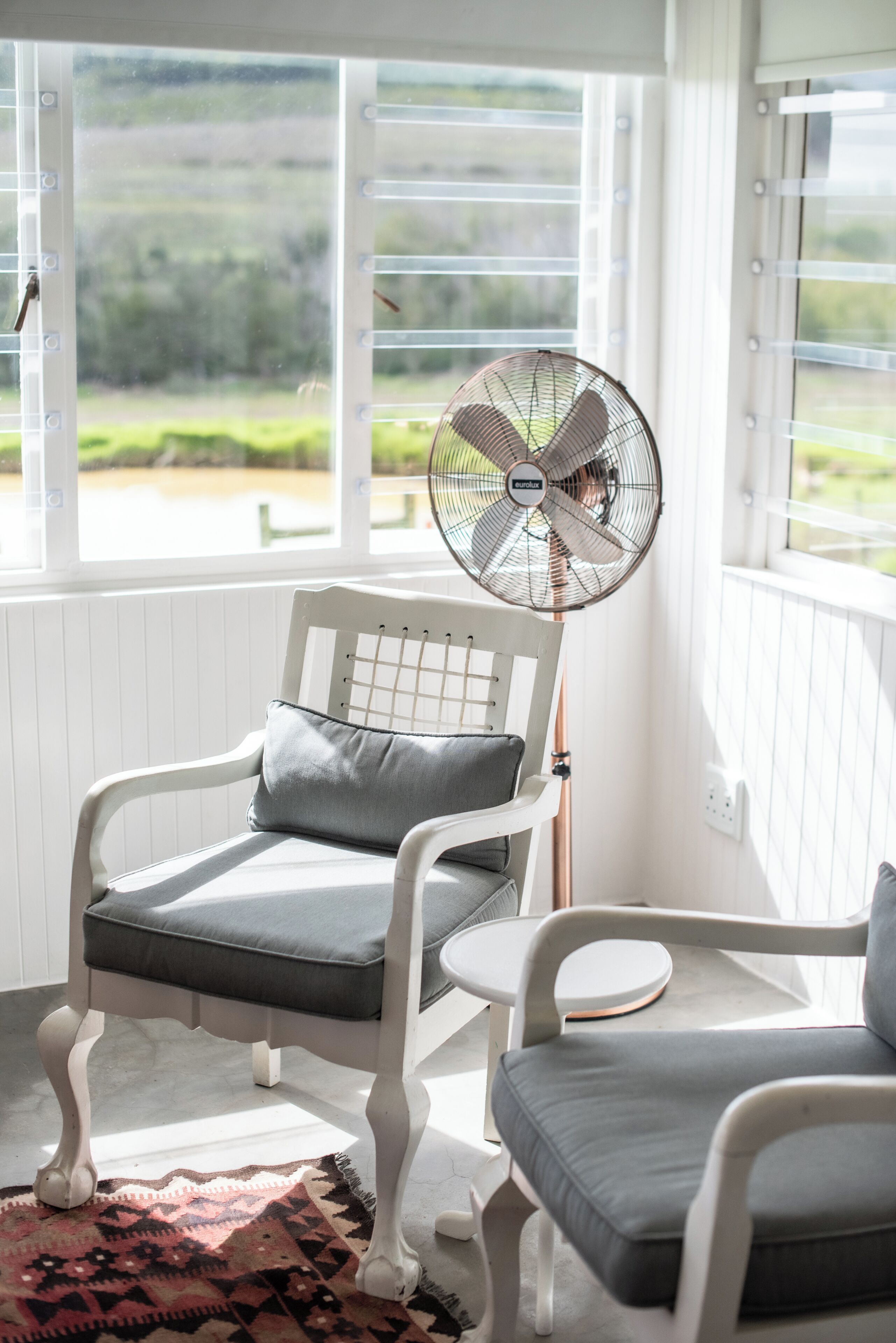 Una silla blanca con un cojín gris y un ventilador de pie en una habitación con paredes blancas y ventanas amplias.