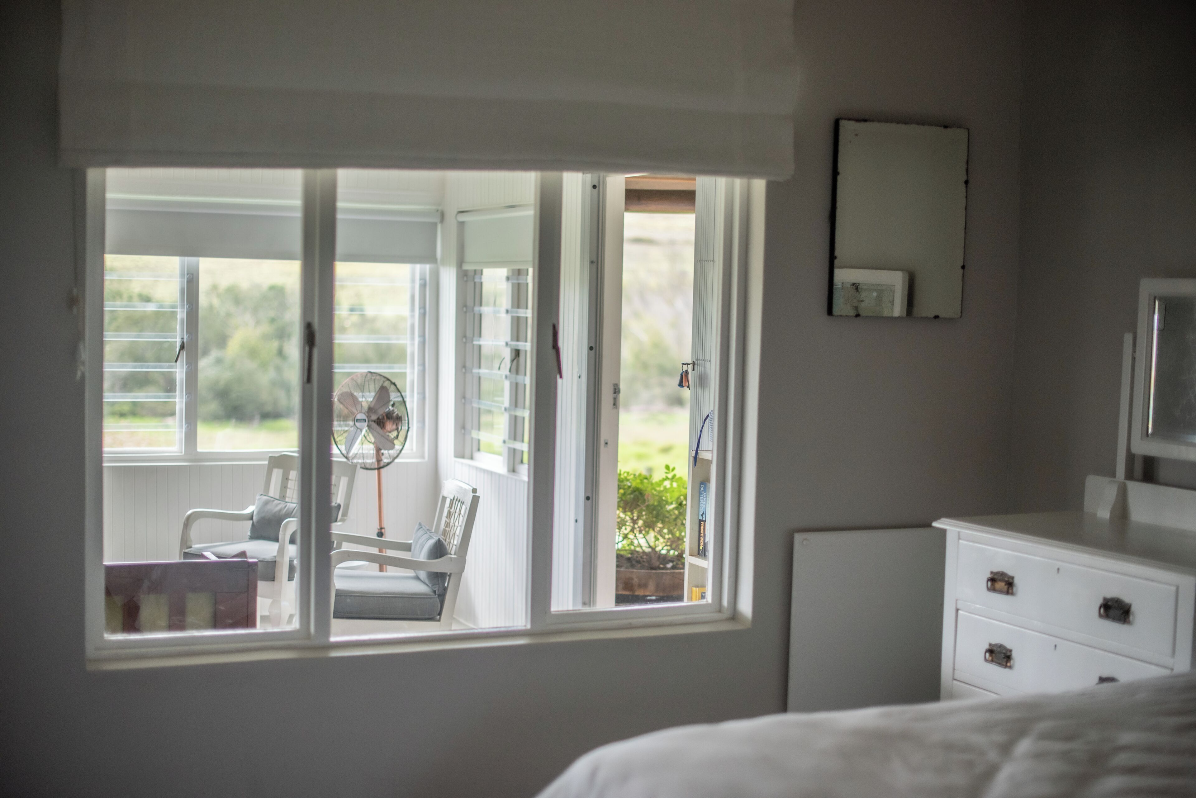 Een slaapkamer met een groot raam, een ventilator, een stoel, een nachtkastje en een dressoir.