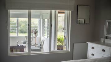 Een slaapkamer met een groot raam, een ventilator, een stoel, een nachtkastje en een dressoir.