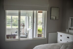 Een slaapkamer met een groot raam, een ventilator, een stoel, een nachtkastje en een dressoir.