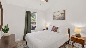 A bedroom with a bed, two bedside tables, a ceiling fan, a window with curtains, a painting on the wall, and a potted plant.