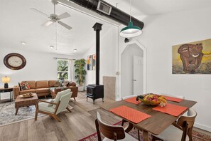 A modern living room with a sofa, chairs, a dining table with a fruit bowl, a fireplace, and a ceiling fan. - Dripping Springs The Wrangler | *NEW* Hill Country Escape (Dripping Springs)