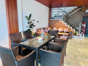 Un moderno vestíbulo de hotel con una mesa de vidrio, sillas de mimbre, un sofá con cojines, una planta en maceta y una escalera con barandilla.