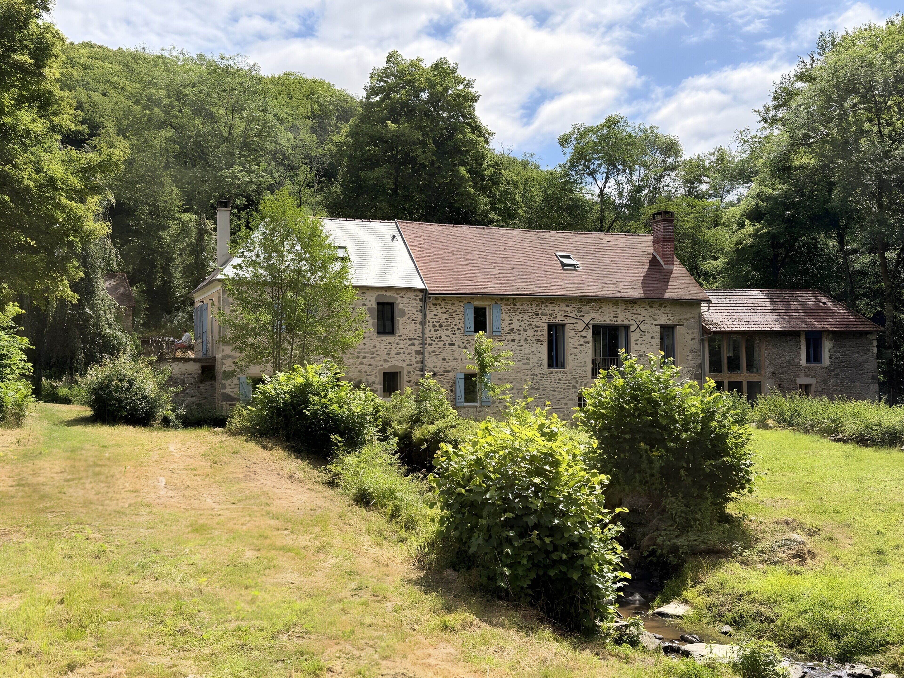 A stone house with a sloped roof, surrounded by lush greenery and a small stream.