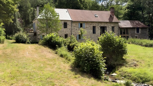 A stone house with a sloped roof, surrounded by lush greenery and a small stream.