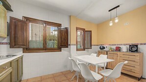 Una cocina con armarios blancos, una mesa blanca y sillas, un ventanal con contraventanas de madera y un suelo de baldosas.