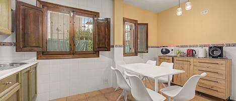 A kitchen with white cabinets, a white table, and chairs, a window with wooden shutters, and a tiled floor.