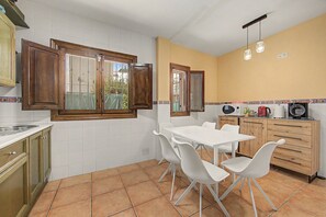 Una cucina con mobili bianchi, un tavolo e sedie bianche, una finestra con persiane in legno e un pavimento piastrellato.