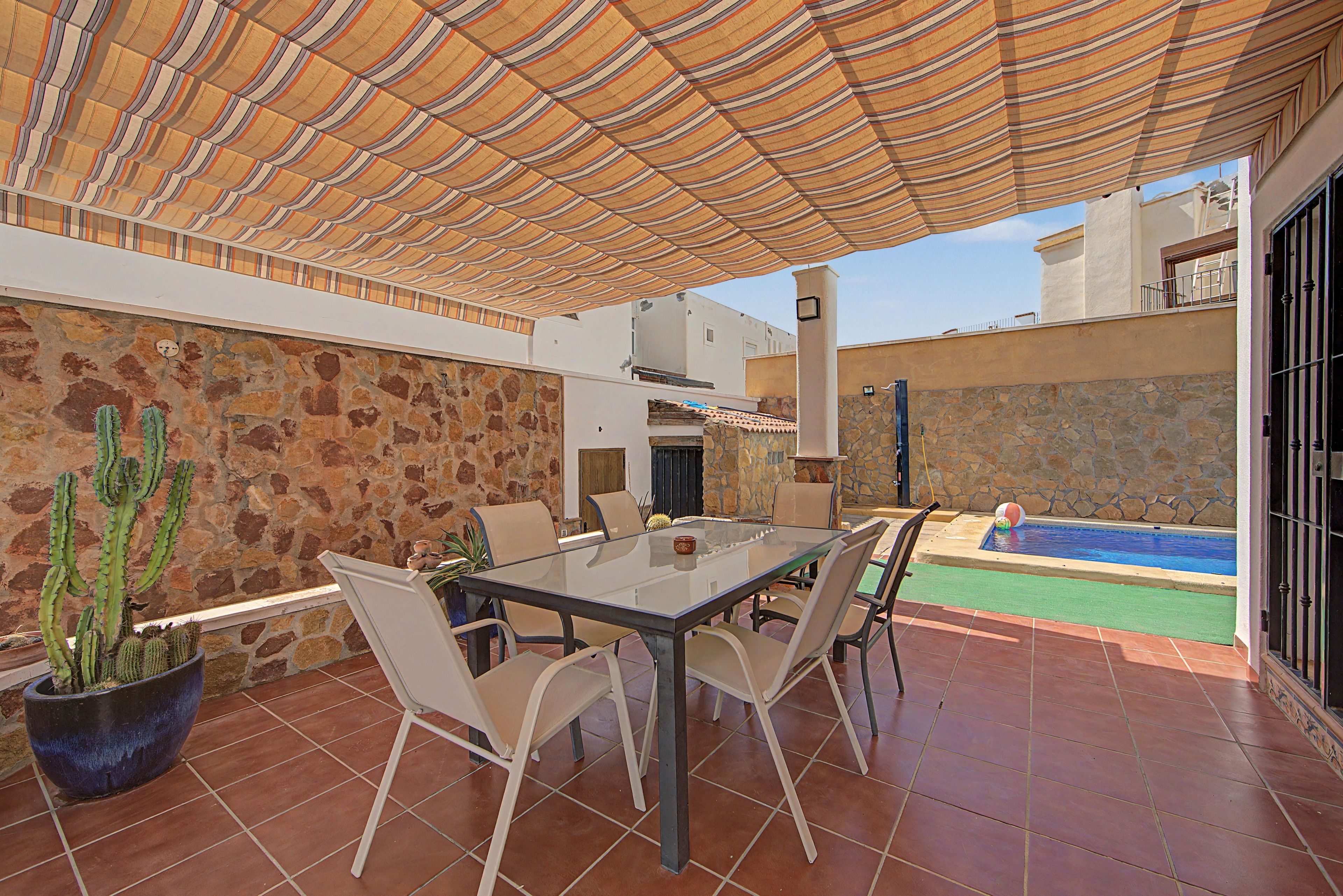 An outdoor patio with a table and chairs, a stone wall, a cactus, and a swimming pool.