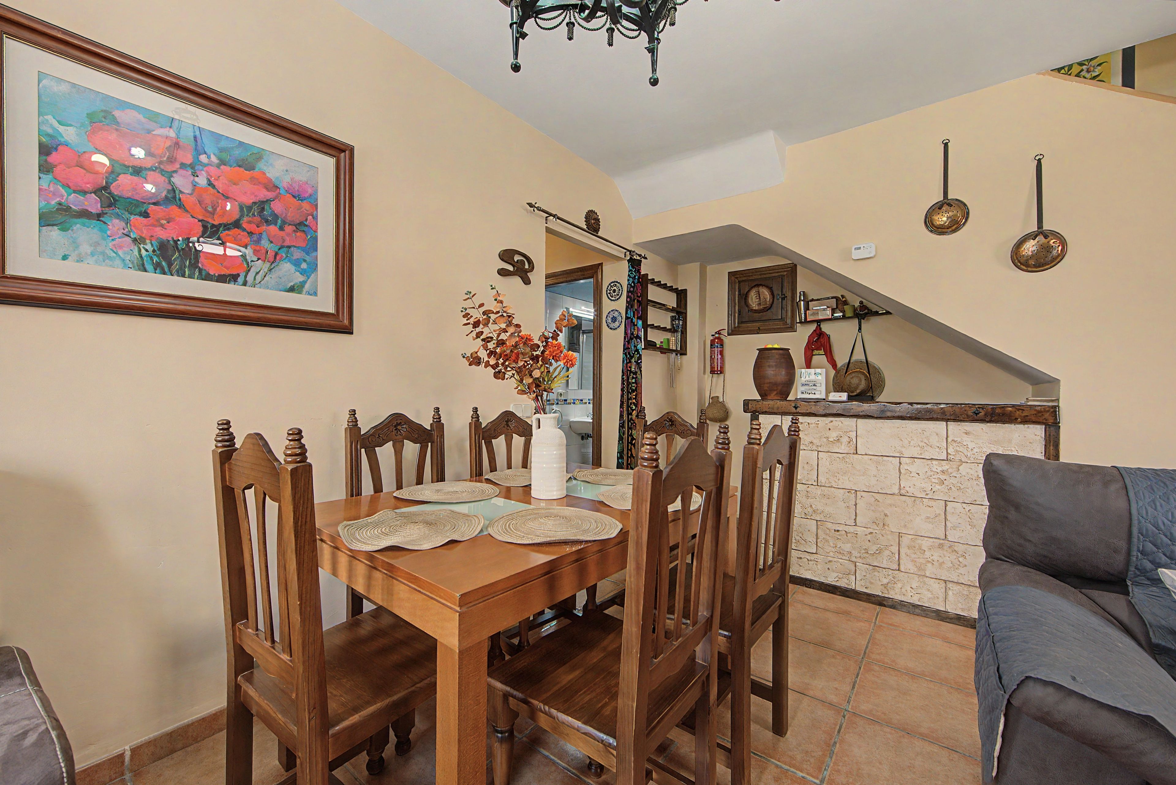 A dining area with a wooden table and chairs, a painting on the wall, and a kitchen in the background.