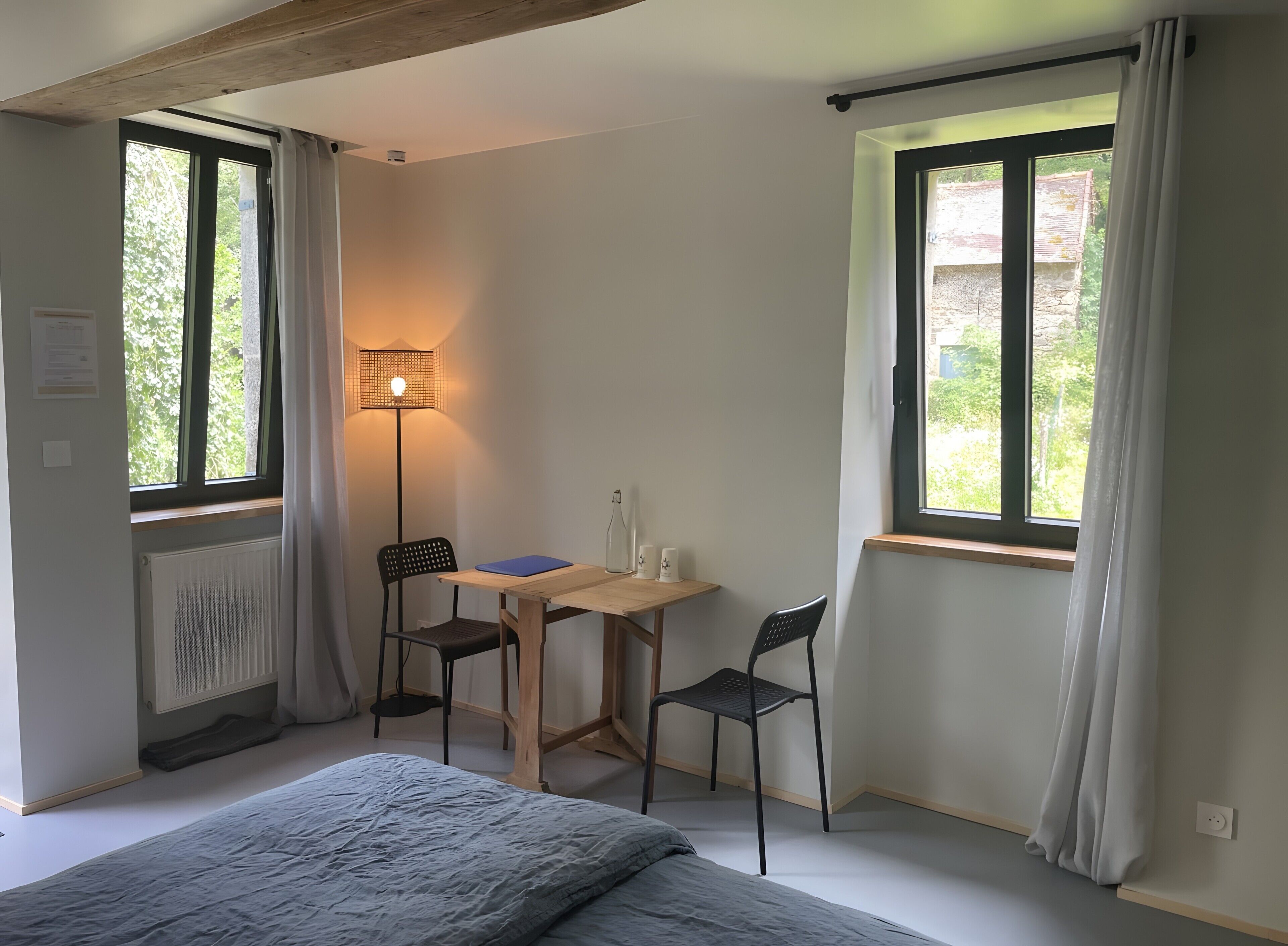Une pièce comprenant un lit, deux chaises, une table, une lampe et deux fenêtres avec des rideaux.