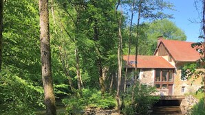 Ein rustikales Steinhaus mit rotem Dach, umgeben von üppigem Grün und einem ruhigen Fluss.