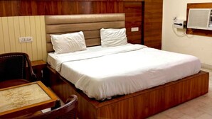 A bedroom with a wooden headboard, a bed with white bedding, a desk with a chair, and a ceiling fan. - Shivmurti Grand  (Haridwar)