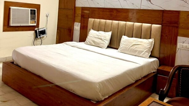 A bedroom with a wooden bed frame, a headboard with built-in lighting, a bedside table, and a ceiling fan. - Shivmurti Grand  (Haridwar)