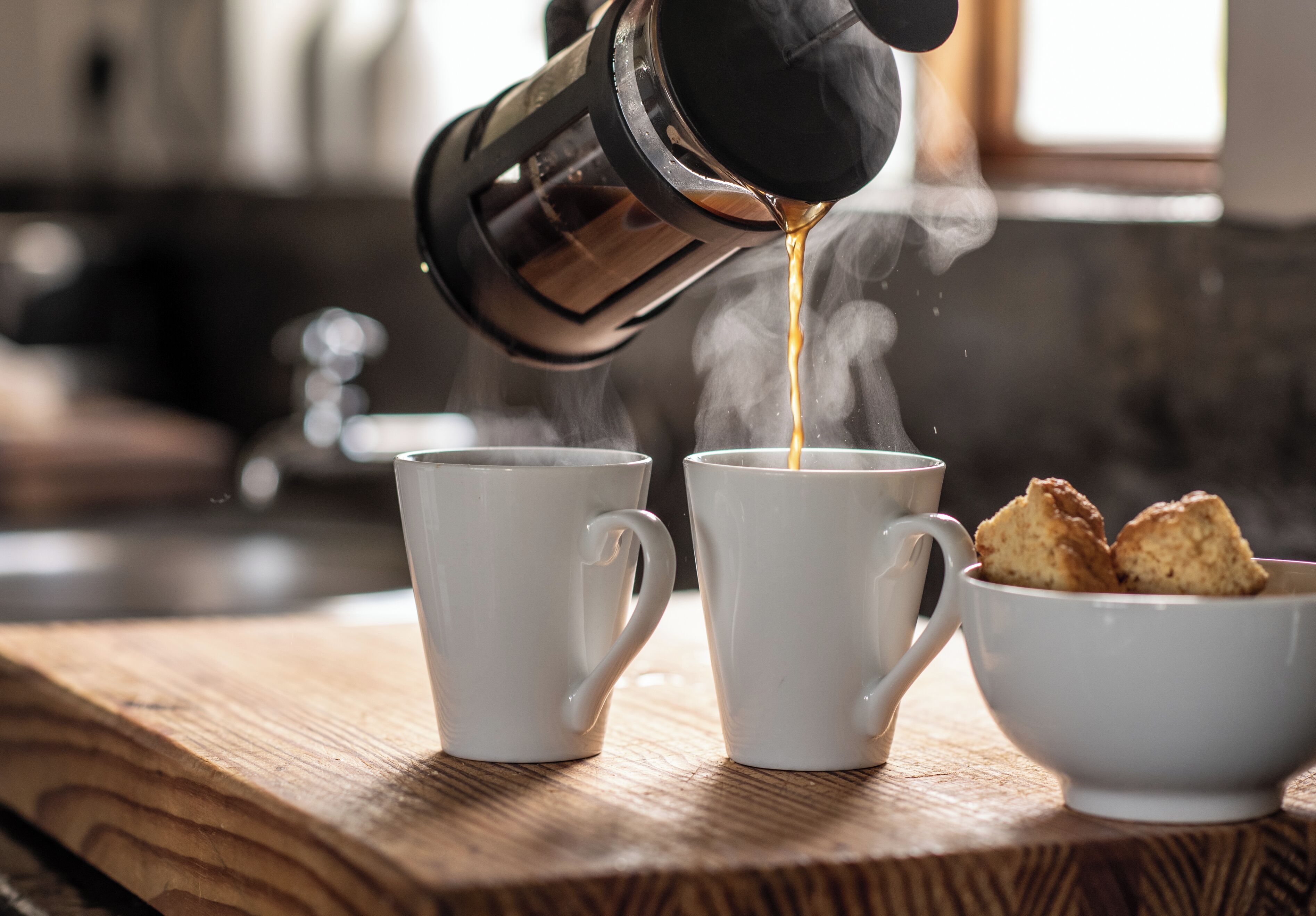 Two white cups on a wooden surface with coffee being poured into one and biscuits in a small bowl.