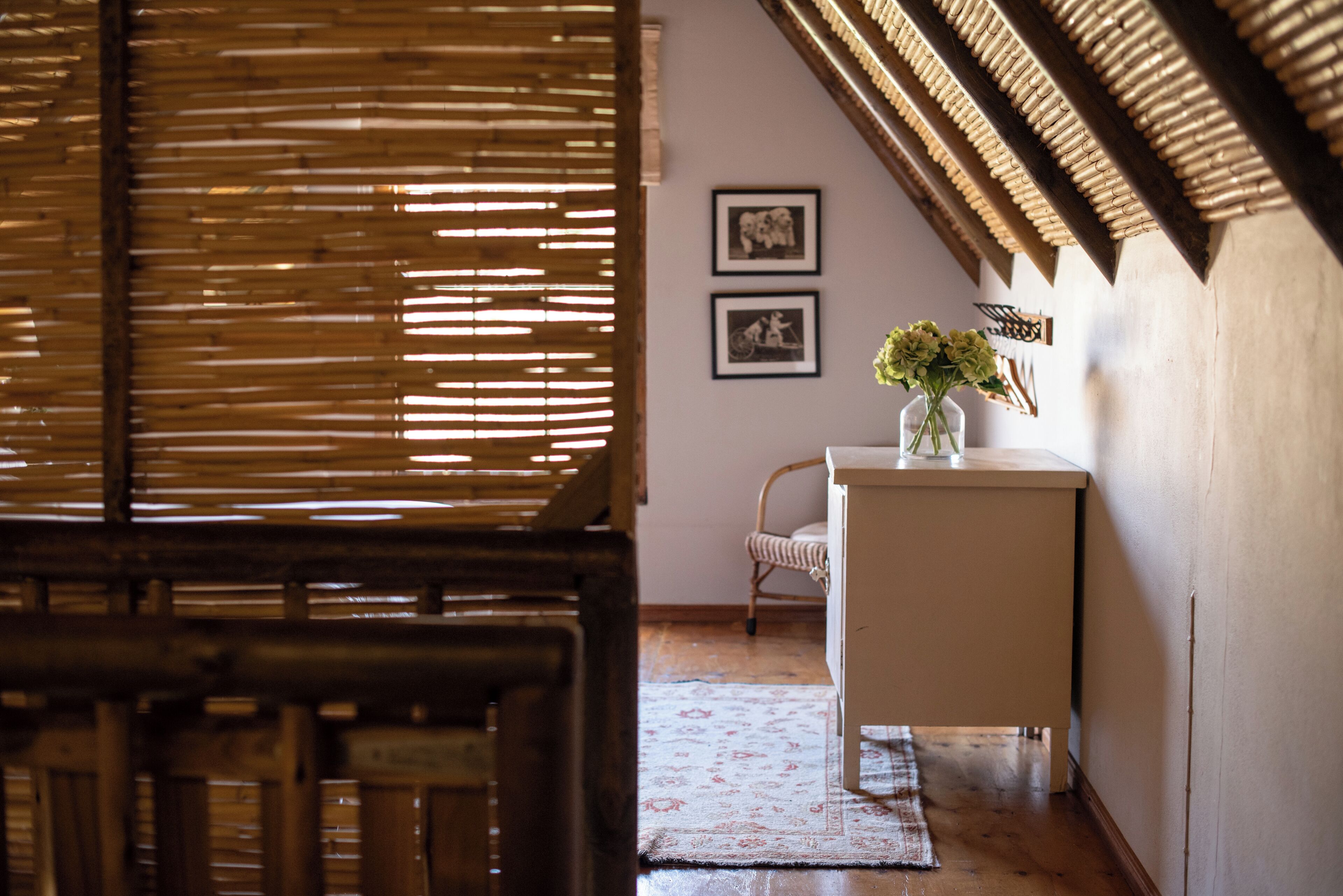 Una habitación con una ventana de listones de madera, una mesa blanca con un florero y dos cuadros en la pared.
