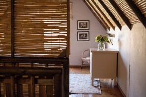 Una habitación con una ventana de listones de madera, una mesa blanca con un florero y dos cuadros en la pared.