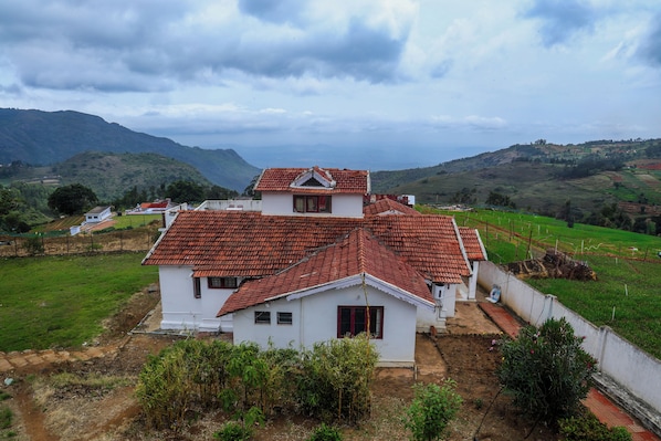 A two-story house with a red-tiled roof, surrounded by green fields and mountains in the background. - Mistora Valley (Ooty)