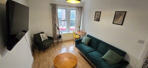 Ein Wohnzimmer mit einem grünen Sofa, einem kleinen runden Holztisch und einem an der Wand befestigten Fernseher.
