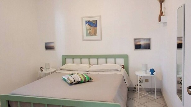 A bedroom with a bed, a bedside table, a chair, a mirror, and a wall-mounted shelf with a plant. - The Residences 3 (Marina di Modica)