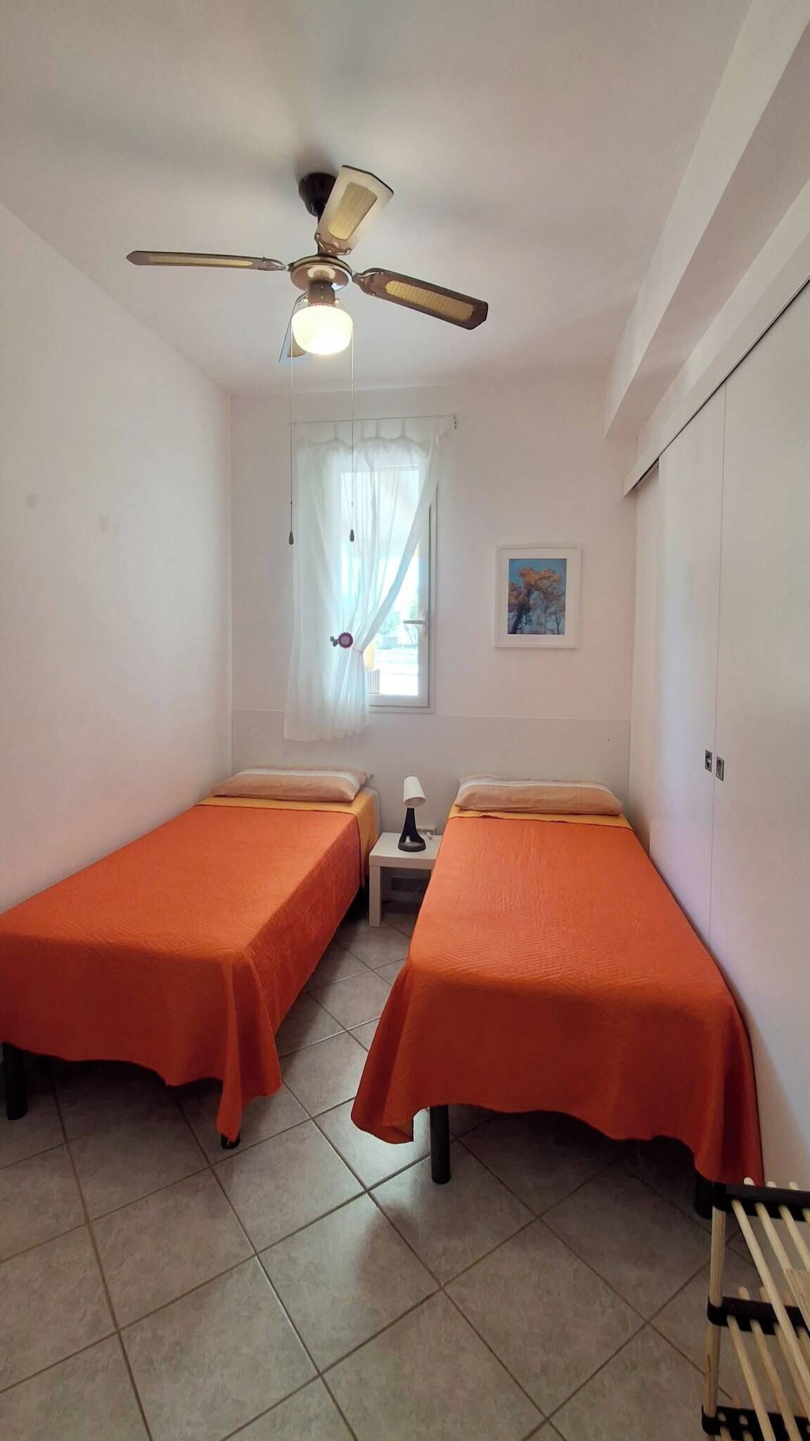 A small, clean room with two single beds, a ceiling fan, a window with curtains, and a small table with a lamp.