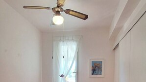 A small, clean room with two single beds, a ceiling fan, a window with curtains, and a small table with a lamp. - The Residences 3 (Marina di Modica)