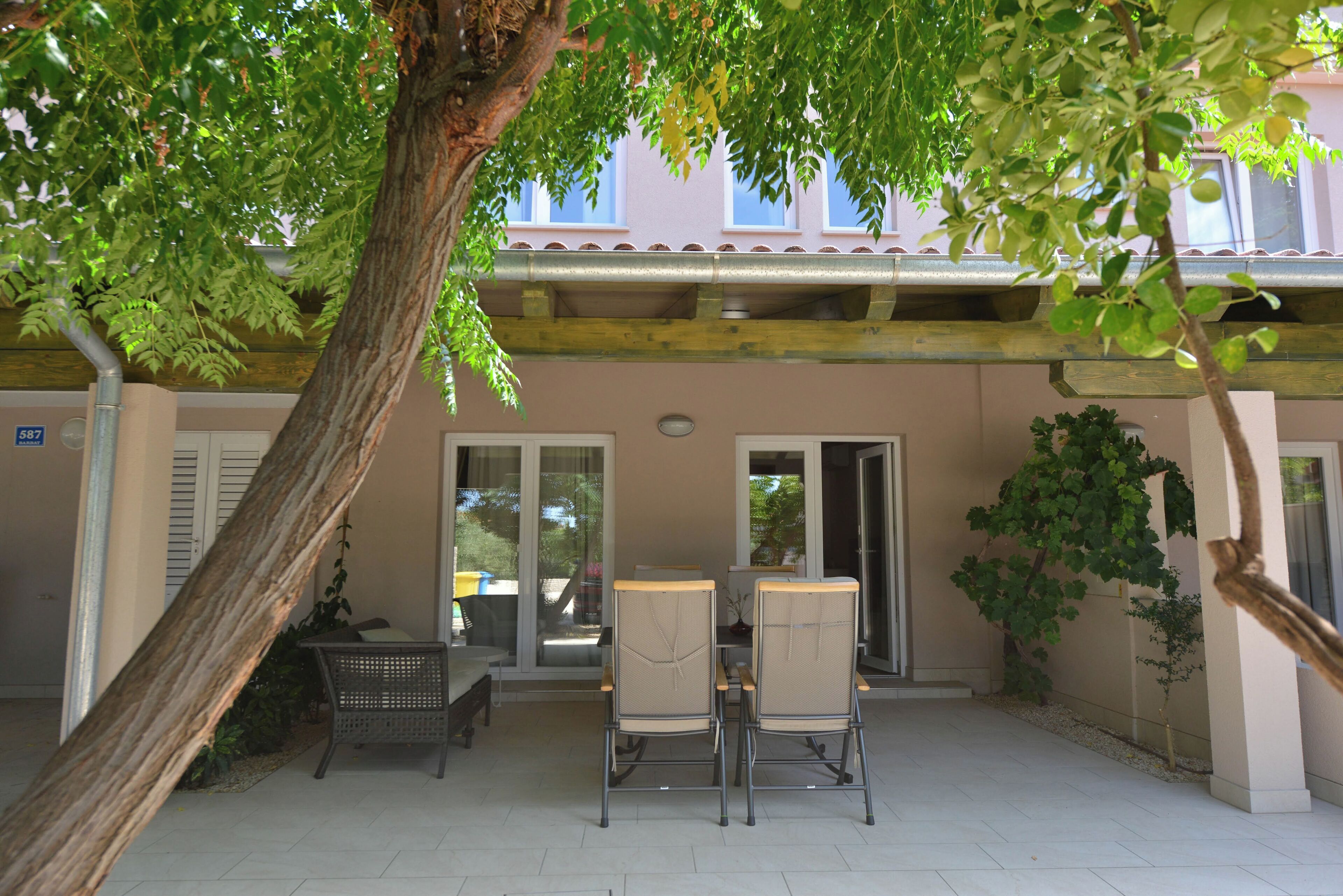 An outdoor seating area with two chairs and a table, surrounded by trees and a building with a tiled roof.