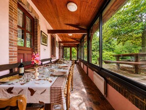 A covered outdoor dining area with brick walls, wooden beams, and large windows overlooking a lush garden. - Family home in a green setting with a large, wooded lot (Rai)