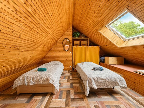 A cozy attic bedroom with wooden walls, two beds, a window, and a small desk area. - Family home in a green setting with a large, wooded lot (Rai)