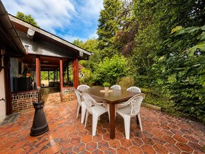 An outdoor patio with a brick floor, a wooden table, and white plastic chairs. - Family home in a green setting with a large, wooded lot (Rai)