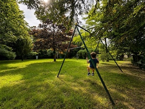 A child on a swing in a grassy park area with trees. - Family home in a green setting with a large, wooded lot (Rai)