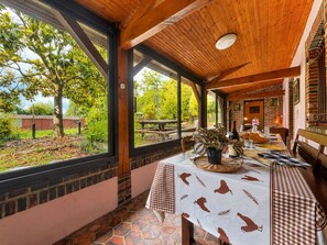 A covered outdoor dining area with a table set for two, a brick wall, and a view of a garden. - Family home in a green setting with a large, wooded lot (Rai)