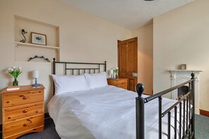A bedroom with a bed, wooden dresser, bedside tables, a nightstand, a wall-mounted shelf with a framed picture, a wooden door, and a fireplace with a mantel. - Family Home With Sea Views (Cromer)
