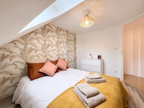 A bedroom with a sloped ceiling, a bed with white bedding and two pillows, a bedside table with a lamp, and a wall with a palm leaf pattern.