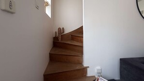 Una escalera con escalones de madera que ascienden, un interruptor de luz en la pared y un pequeño ventanal que deja entrar luz natural.