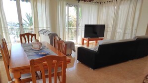 A dining area with a table and chairs, a sofa, a television, and a view of the outdoors through large windows. - Beachfront Apartment (S'Arenal)