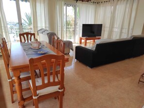Ein Essbereich mit Tisch und Stühlen, einer Couch, einem Fernseher und Blick ins Freie durch große Fenster.