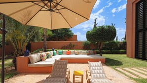 Ein sonniger Sitzbereich im Freien mit einem großen Sonnenschirm, einer gepolsterten Couch mit bunten Kissen und zwei Liegestühlen mit Handtüchern.