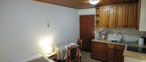 Une pièce de taille modeste comprenant un lit, une table à manger avec des chaises, un coin cuisine avec des placards en bois et un sol carrelé.