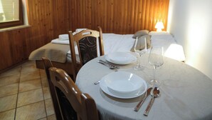 Een eetgedeelte met een ronde tafel gedekt voor twee personen, een bed met houten hoofdbord op de achtergrond en een raam met blinds.
