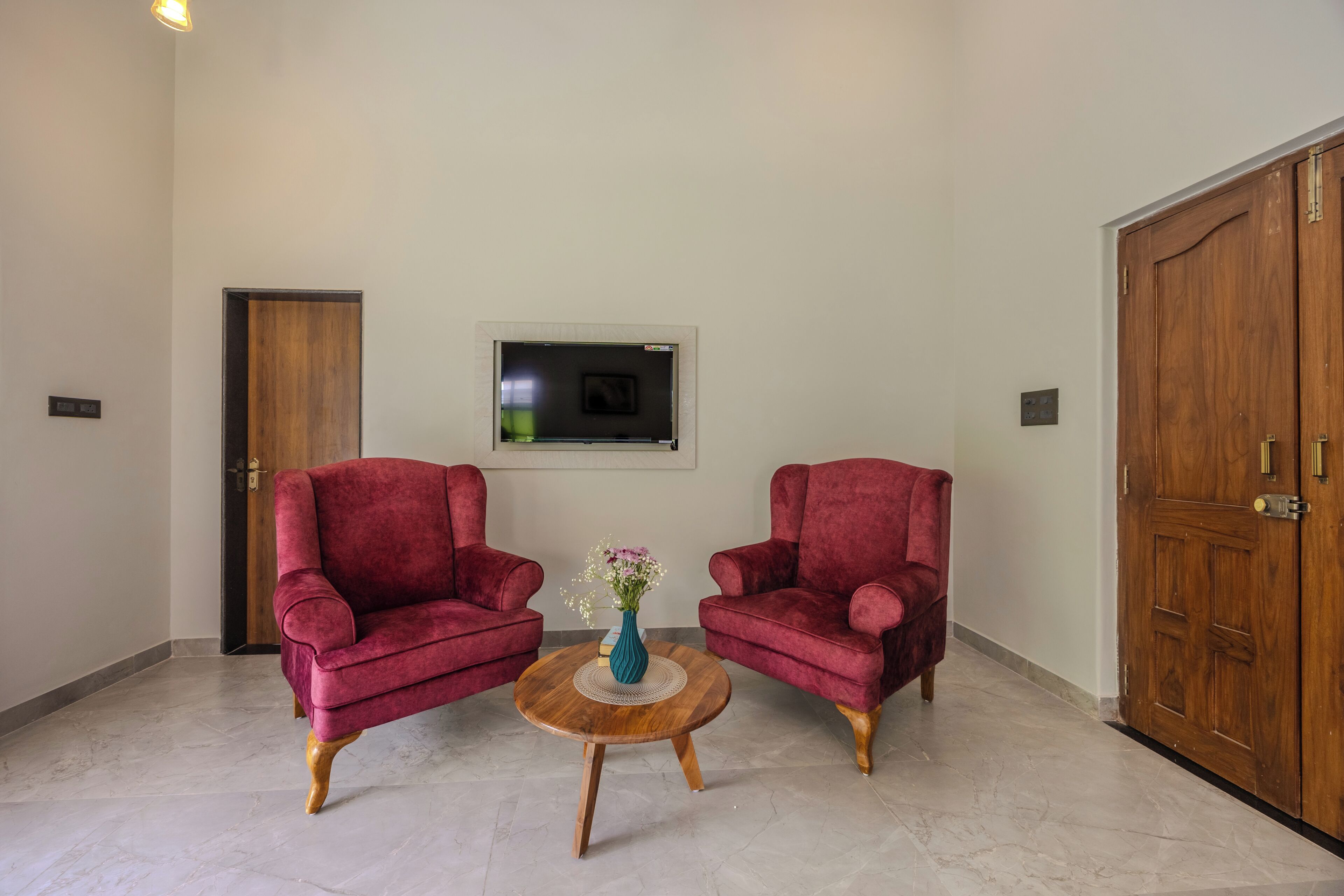 A room with two red armchairs, a small wooden table with a vase, a door, and a wall-mounted television.