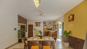 A dining area with a table set for four, chairs, a ceiling fan, and a kitchen visible in the background. - ELIVAAS Casa Vilson 3BHK Villa with Pool (Salvador du Mundu)