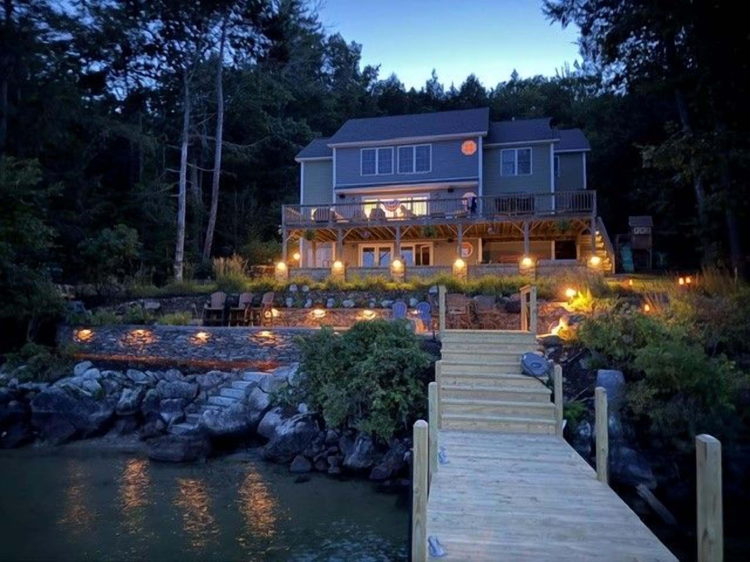 A two-story house with a dock leading to a body of water, surrounded by trees.