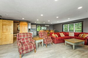 A spacious living area with a red sofa set, a wooden coffee table, and plaid armchairs. A kitchen is visible in the background with a dining area. - NEW FOR 2027 Lake Winnipesaukee Waterfront Sleeps 16 (Moultonborough)